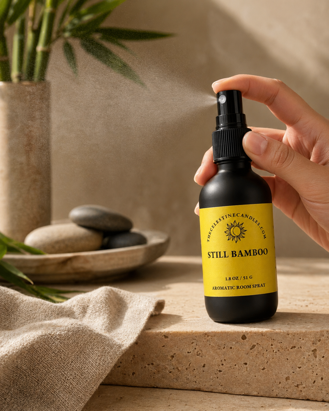 Hand holding a 'Still Bamboo' aromatic room spray bottle with a plant and stones in the background.